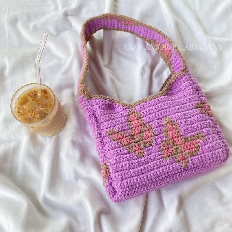 May include: A purple crocheted tote bag with a brown and pink floral design. The bag has a long strap and the text "CON AMOR BY ARTISANS" is visible on the strap.