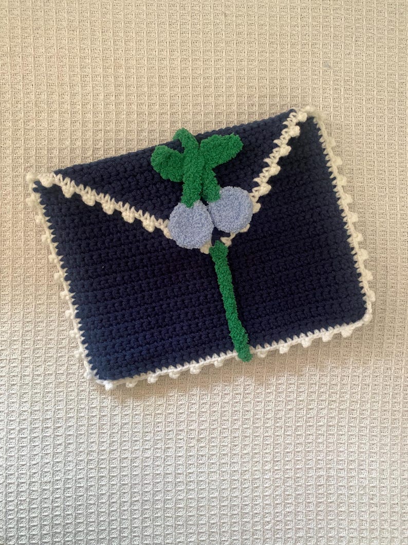 Puede incluir: Un embrague de sobre de crochet azul con un ribete de crochet blanco y una decoraci&oacute;n de flor de fieltro verde y azul.
