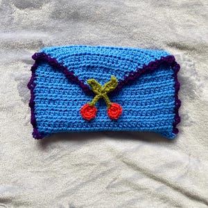 Puede incluir: Un embrague de crochet azul con un borde morado y un dise&ntilde;o de cerezas. Las cerezas son rojas con tallos y hojas verdes.