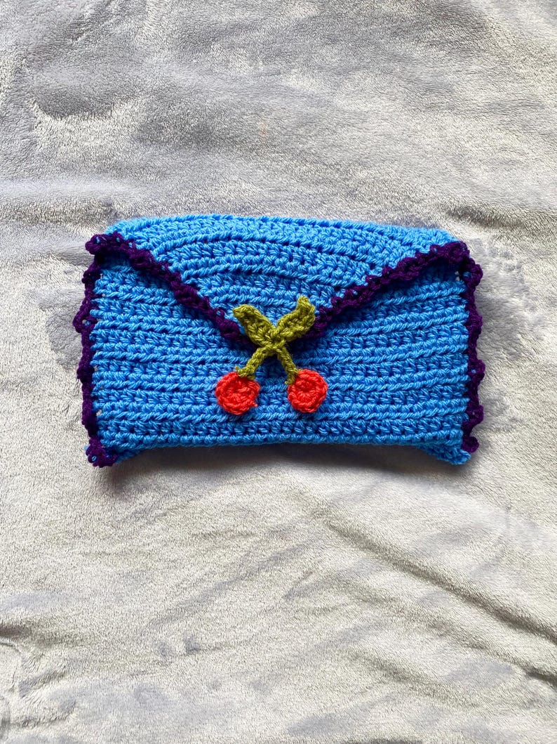 Puede incluir: Un embrague de crochet azul con un borde morado y un dise&ntilde;o de cerezas. Las cerezas son rojas con tallos y hojas verdes.