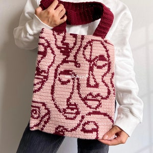 K&ouml;nnte beinhalten: Eine geh&auml;kelte Tragetasche in Rosa und Burgunderrot. Die Tasche hat ein sich wiederholendes Muster aus abstrakten Gesichtern.