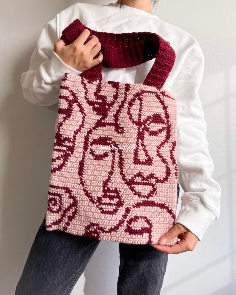 K&ouml;nnte beinhalten: Eine geh&auml;kelte Tragetasche in Rosa und Burgunderrot. Die Tasche hat ein sich wiederholendes Muster aus abstrakten Gesichtern.