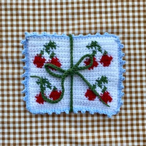 Puede incluir: Un cuadrado tejido a crochet con un borde azul y una cinta verde atada en un lazo. El cuadrado est&aacute; decorado con cerezas rojas y verdes.