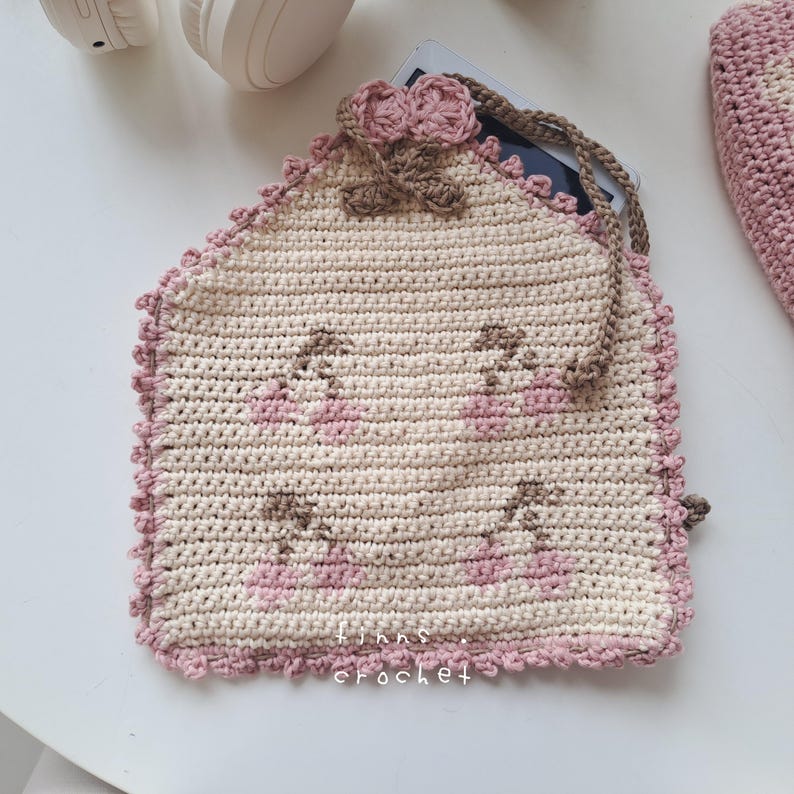 Puede incluir: Una bolsa de crochet con un dise&ntilde;o de cerezas rosa y marr&oacute;n. La bolsa tiene un borde de crochet rosa y marr&oacute;n y una correa de crochet marr&oacute;n. La bolsa est&aacute; hecha por "finns crochet".