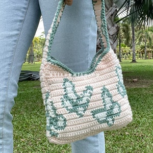 May include: A crocheted handbag with a light pink base and green leaf pattern. The bag has a single strap and is being held by a person wearing blue jeans.