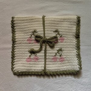Puede incluir: Un cuadrado blanco tejido a crochet con un borde verde y un lazo rosa y verde en el centro. El cuadrado tiene un patr&oacute;n de flores rosas con tallos verdes.