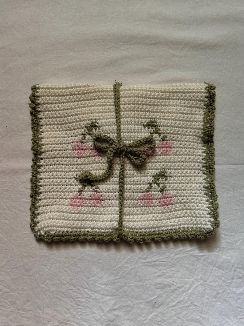 Puede incluir: Un cuadrado blanco tejido a crochet con un borde verde y un lazo rosa y verde en el centro. El cuadrado tiene un patr&oacute;n de flores rosas con tallos verdes.