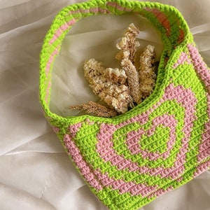 May include: A green and pink crocheted tote bag with a single strap. The bag is made with a repeating pattern of squares and circles. The bag is empty and sitting on a white surface.
