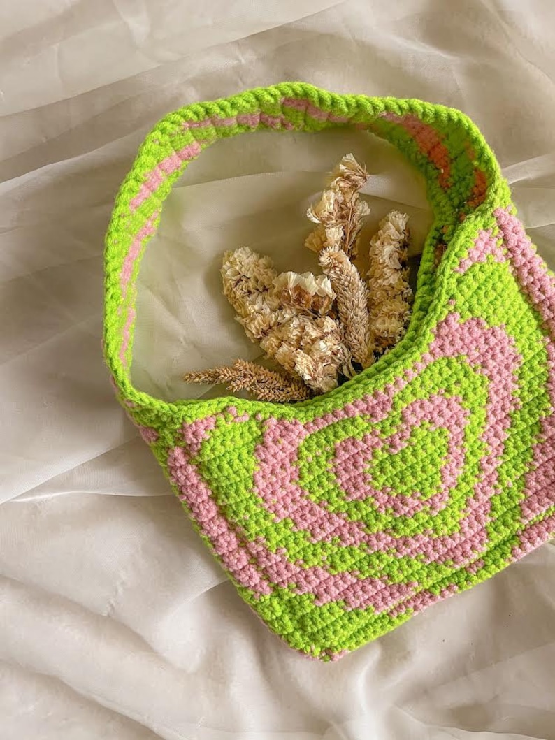 May include: A green and pink crocheted tote bag with a single strap. The bag is made with a repeating pattern of squares and circles. The bag is empty and sitting on a white surface.