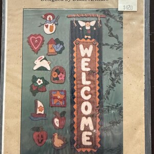May include: A craft kit titled "Month by Month Welcome" designed by Diane Arthurs. The kit includes a welcome sign and various felt appliques, such as a snowman, heart, birdhouse, rabbit, and apple. The sign reads "WELCOME".