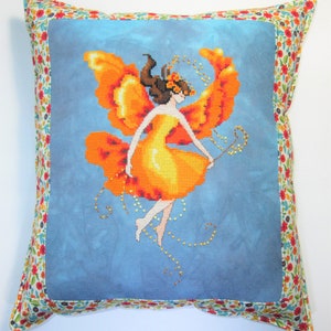 May include: A decorative pillow with a blue background and a cross-stitch design of a fairy with orange wings and a yellow dress. The pillow has a floral patterned border.