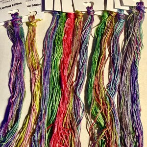 May include: A collection of colorful silk embroidery floss in shades of purple, green, red, and yellow. Each skein is labeled "Ship's Manor Silk, 100% French Spun Silk, 12 Strand, 5 Yards, Limited Edition."