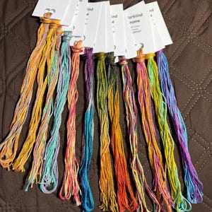 May include: A collection of colorful embroidery floss skeins, each with a white tag. The floss features a variety of blended colors, including yellow, orange, pink, blue, and green. The tags read "Colorblind Gnome" and include details about the floss.