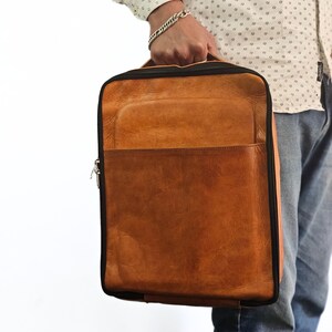May include: A brown leather backpack with a black zipper and trim. The backpack has a large front pocket and a smaller pocket on the top.