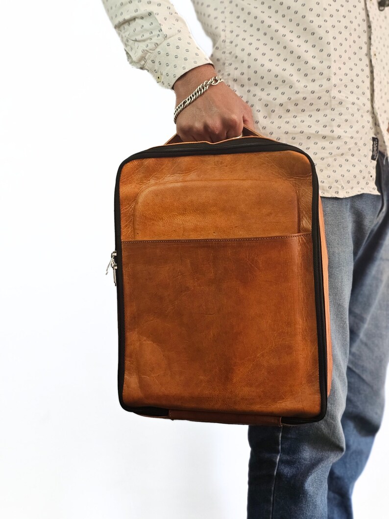 May include: A brown leather backpack with a black zipper and trim. The backpack has a large front pocket and a smaller pocket on the top.