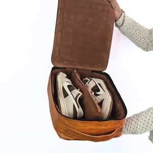 May include: A brown leather shoe bag with a zip closure and a handle. The bag is open and two white and brown trainers are inside.