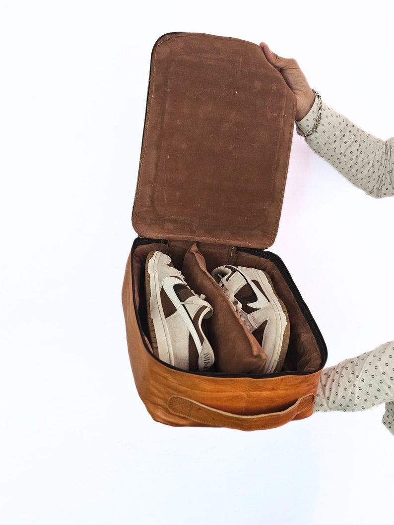 May include: A brown leather shoe bag with a zip closure and a handle. The bag is open and two white and brown trainers are inside.