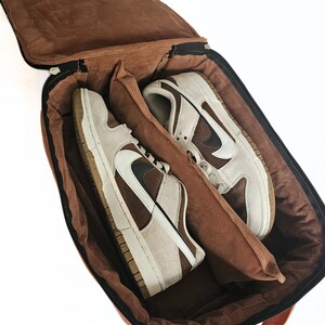 May include: A brown leather shoe bag with a zip closure. The bag is open and two pairs of brown and white trainers are inside.