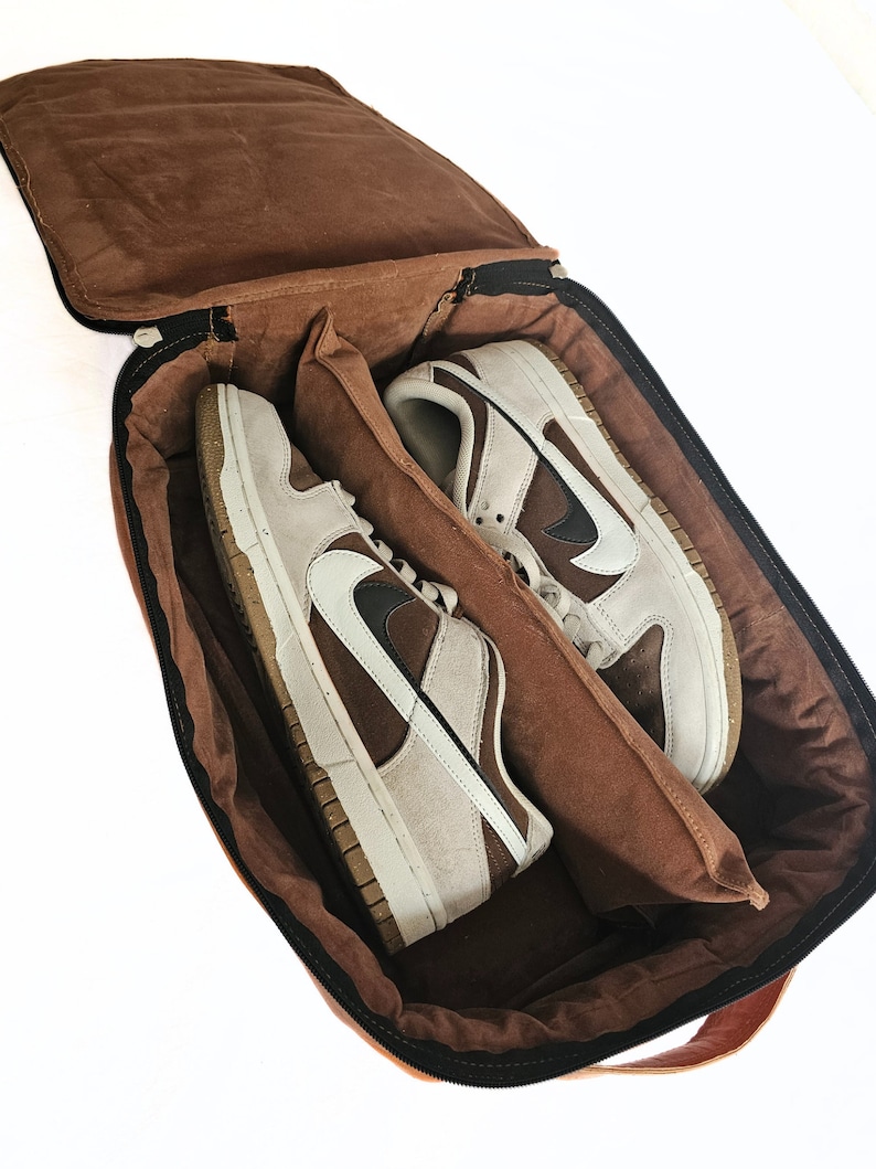 May include: A brown leather shoe bag with a zip closure. The bag is open and two pairs of brown and white trainers are inside.