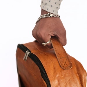 May include: A brown leather backpack with a black zipper and a strap. The backpack is being held by a hand wearing a silver bracelet.