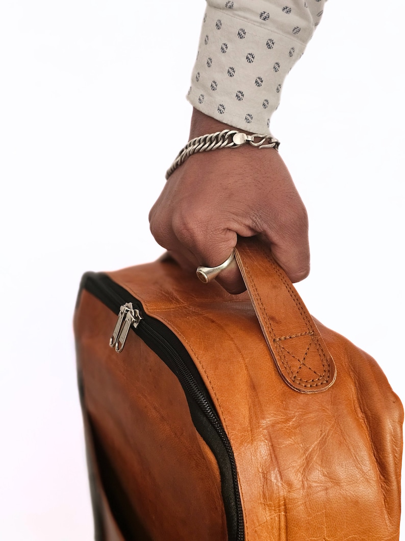 May include: A brown leather backpack with a black zipper and a strap. The backpack is being held by a hand wearing a silver bracelet.