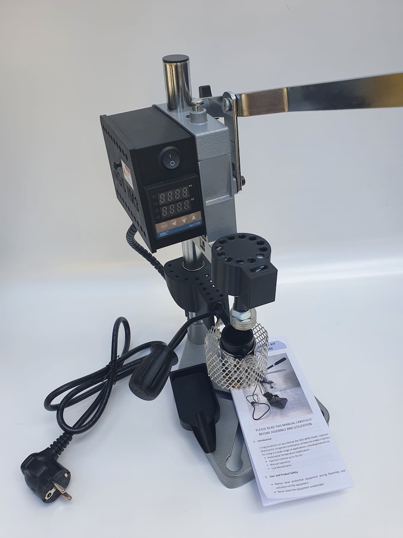 May include: A silver and black heat press machine with a digital temperature display. The machine has a lever and a power cord. The machine is on a white surface. The text on the instruction manual reads "PLEASE READ THIS MANUAL CAREFULLY BEFORE ASSEMBLY AND UTILIZATION."