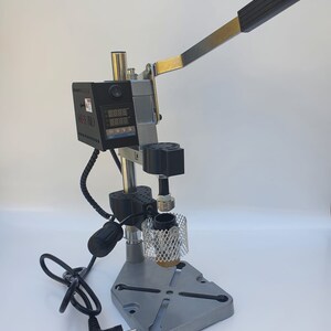 May include: A silver and black metal press with a digital display and a lever. The press has a black power cord and a black plug. The press is on a white surface.