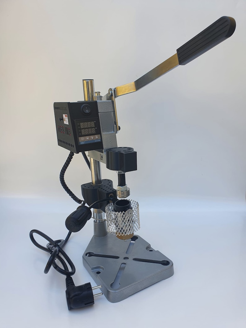 May include: A silver and black metal press with a digital display and a lever. The press has a black power cord and a black plug. The press is on a white surface.
