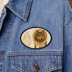 May include: Oval patch featuring a wolf's head within a dreamcatcher design, with feathers and geometric patterns. The patch is set against a cream background with a black border, and is affixed to a denim jacket.