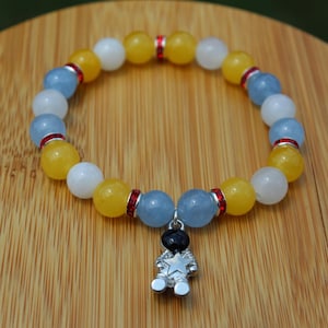 May include: A beaded stretch bracelet featuring alternating round yellow, white, and light blue beads. The bracelet is accented with small, cylindrical red spacers and features a silver-toned metal charm shaped like an astronaut holding a star.