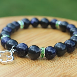May include: A beaded bracelet featuring a mix of dark blue and black beads. The bracelet includes a silver cloud charm and green rhinestone accents. The beads have a speckled, glittery appearance. The bracelet is displayed on a wooden surface.