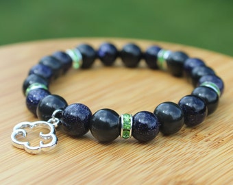 Pode incluir: Pulseira de contas com contas azuis escuras e pretas, um berloque de nuvem prateada e detalhes de strass verdes. As contas têm uma aparência salpicada e brilhante. A pulseira é exibida em uma superfície de madeira. Circunferência de aproximadamente 19 cm.