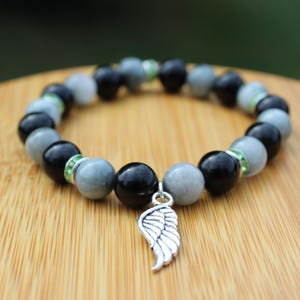 Puede incluir: Una pulsera de cuentas redondas negras y grises, acentuada con espaciadores de pedrería verdes. Un dije de ala plateada cuelga de la pulsera. La pulsera se muestra sobre una superficie de madera.