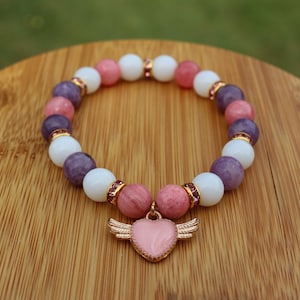 May include: A pink and purple gemstone bracelet with a pink heart charm with gold wings. The bracelet is made with white, pink, and purple beads.