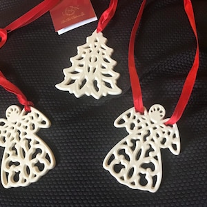 May include: Three white porcelain Christmas ornaments with red ribbon. The ornaments are a tree, an angel, and another angel. The tree ornament has a lacy design. The angel ornaments have lacy wings and a halo.