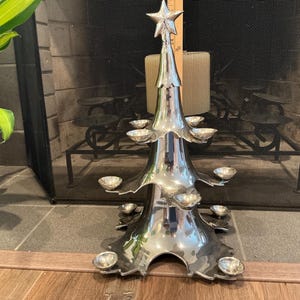 May include: A silver-colored Christmas tree-shaped candle holder with multiple small candle cups. The tree has a star on top and is placed on a wooden floor. The background includes a fireplace screen and a green plant.