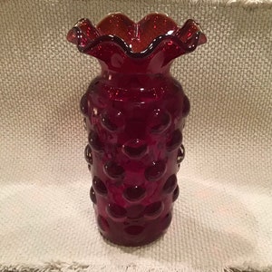 May include: A ruby red glass vase with a ruffled rim and a textured, bubble-like pattern. The vase is cylindrical with a flared top and is approximately 8 inches tall.