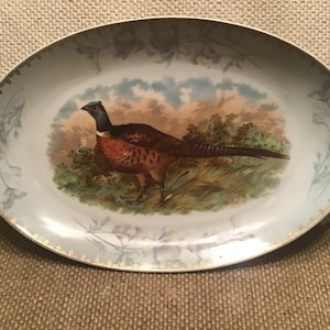 May include: Oval ceramic serving platter with a pheasant illustration. The bird is depicted in a naturalistic setting with brown, orange, and green hues. The rim features a floral design with gold accents.