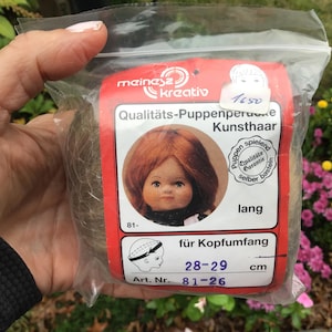 May include: A package of long, light brown doll hair for making dolls. The package includes a doll head with red hair and a label that reads "Qualitäts-Puppenperücke Kunsthaar" and "Art. Nr. 81-26".