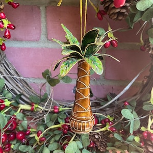 May include: A gold palm tree ornament with green leaves and a gold string for hanging. The ornament is surrounded by a wreath of green leaves and red berries.