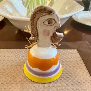 May include: A ceramic bell-shaped figurine with a stylized face and hair. The face is white with brown hair and a pink cheek. The bell has orange, purple, and yellow stripes. Decorative item.