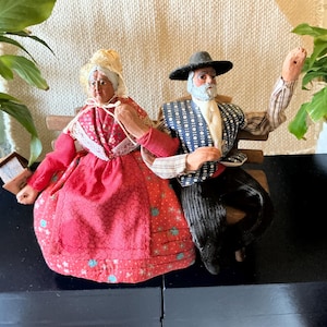 May include: Two vintage dolls dressed in traditional clothing are sitting on a wooden bench. The woman is wearing a red dress with white lace and a white bonnet. The man is wearing a blue and white patterned jacket with a black hat and a white beard.