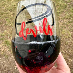 May include: A stemless wine glass with a black and red glitter base. The clear glass has a black hockey puck design and the word "devils" in red script. The glass is held in a hand against a blurred green background.
