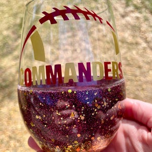 May include: A clear glass wine glass with a burgundy glitter base. The glass features a gold and burgundy football design with the word "COMMANDERS" on the side.