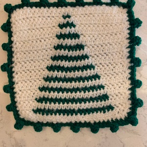 Op de afbeelding: Een wit gebreid vierkant met een groen kerstboomontwerp. Het vierkant heeft een groene gehaakte rand met pompoentjes.