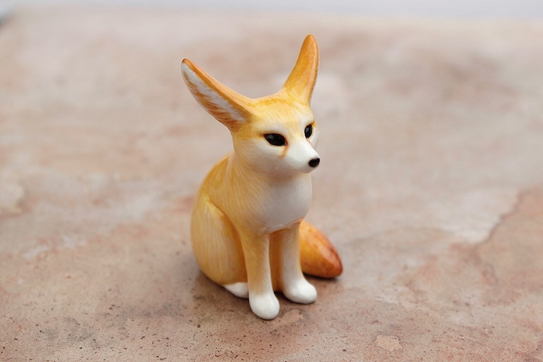 Puede incluir: Una peque&ntilde;a figurita de cer&aacute;mica de un zorro fennec sentado con las orejas levantadas. El zorro es de color marr&oacute;n claro con marcas blancas en la cara y el pecho.