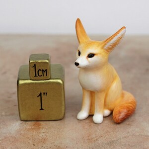 Puede incluir: Una peque&ntilde;a figurita de cer&aacute;mica pintada a mano de un zorro fennec, sentado con las orejas levantadas. La figurita mide aproximadamente 1 pulgada de alto y est&aacute; junto a un cubo de 1 cm para la escala.