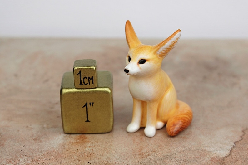 Puede incluir: Una peque&ntilde;a figurita de cer&aacute;mica pintada a mano de un zorro fennec, sentado con las orejas levantadas. La figurita mide aproximadamente 1 pulgada de alto y est&aacute; junto a un cubo de 1 cm para la escala.