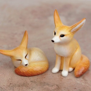 Puede incluir: Dos peque&ntilde;as figuritas de zorro de cer&aacute;mica. Un zorro est&aacute; acurrucado durmiendo, mientras que el otro est&aacute; sentado erguido con las orejas levantadas.