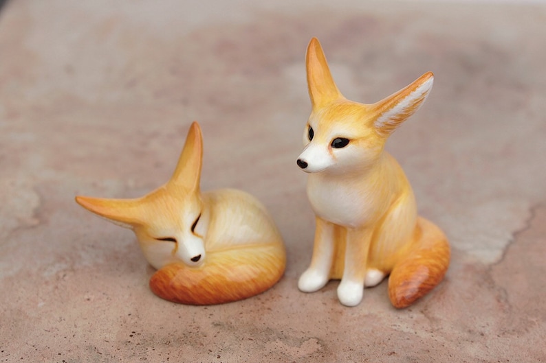 Puede incluir: Dos peque&ntilde;as figuritas de zorro de cer&aacute;mica. Un zorro est&aacute; acurrucado durmiendo, mientras que el otro est&aacute; sentado erguido con las orejas levantadas.